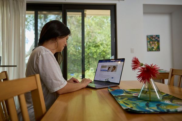 A lady sitting at a wooden dining table, in front of glass doors leading out to a garden. She is visiting the SA Water website, on a laptop, with her fingers above the keypad.